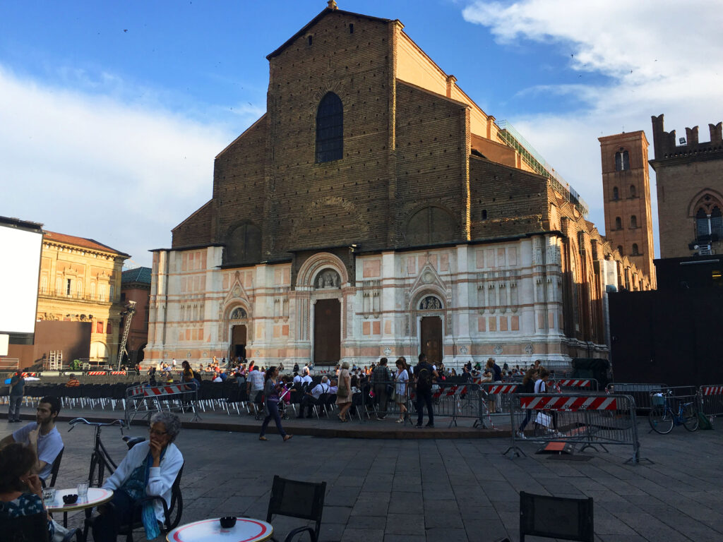 La foto mostra Piazza Maggiore. Davanti alla chiesa di San Petronio sono presenti delle sedie rivolte verso uno schermo, in attesa della proiezione di un film all'aperto